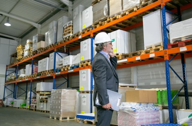 Focused CEO in formal suit and hardhat standing in warehouse Focused CEO in formal suit and hardhat standing in warehouse and talking on mobile phone. Shelves with goods on wooden pallets. Side view. Business or logistics concept